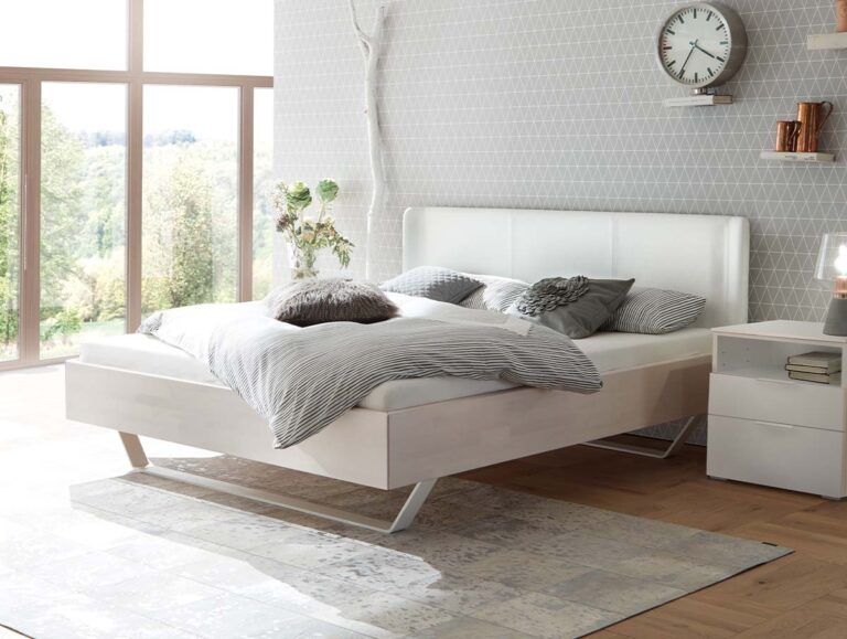 Modernes Schlafzimmer mit einem weißen Bett, grauer und weißer Bettwäsche, einer geometrisch gemusterten Akzentwand, einem großen Fenster mit Blick nach draußen, einem Nachttisch, Dekorationsartikeln und einer Wanduhr. Lichtdurchfluteter, minimalistischer und aufgeräumter Raum.