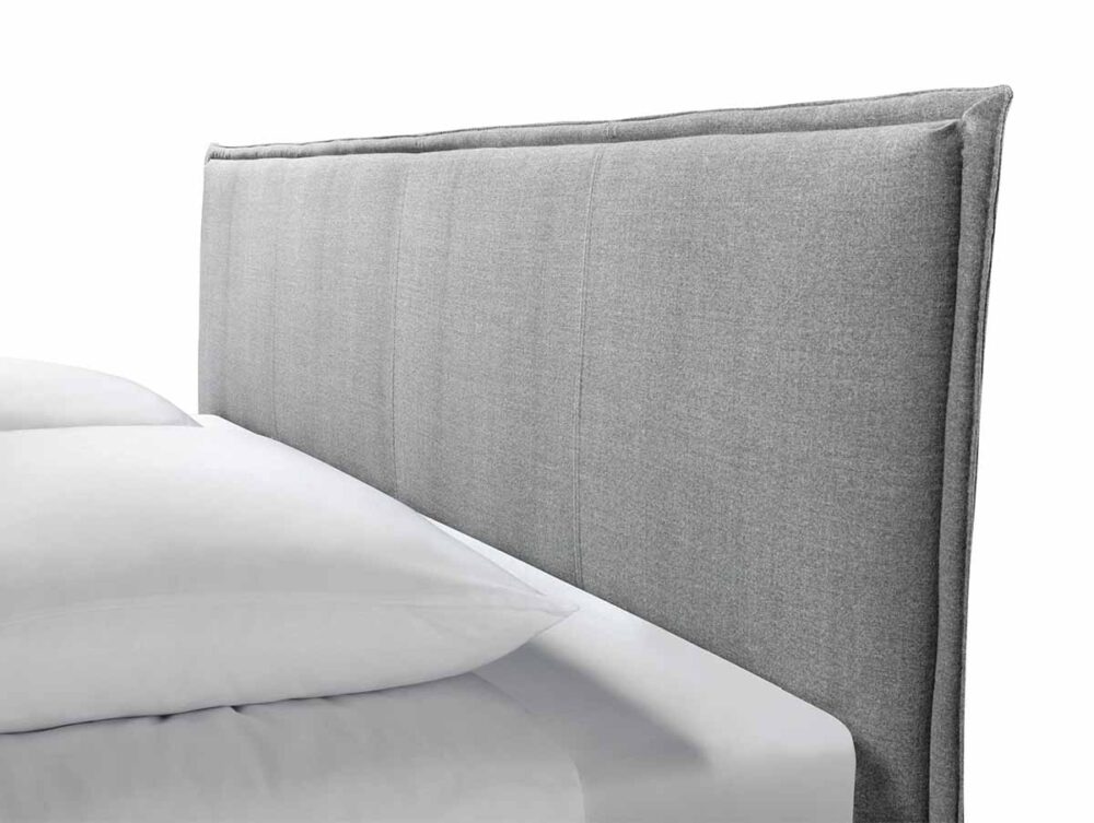 Eine Nahaufnahme eines modernen Bettes mit einem hellgrauen, gepolsterten Stoffkopfteil und weißer Bettwäsche vor einem schlichten weißen Hintergrund.