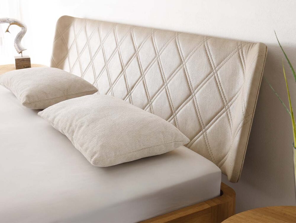 Eine Nahaufnahme eines Bettes mit einem hellbeigen, rautenförmig gepolsterten Kopfteil, zwei passenden Kissen und einem neutralen Bettlaken. Die Ecke eines hölzernen Nachttisches und eine dekorative Pflanze sind teilweise sichtbar.