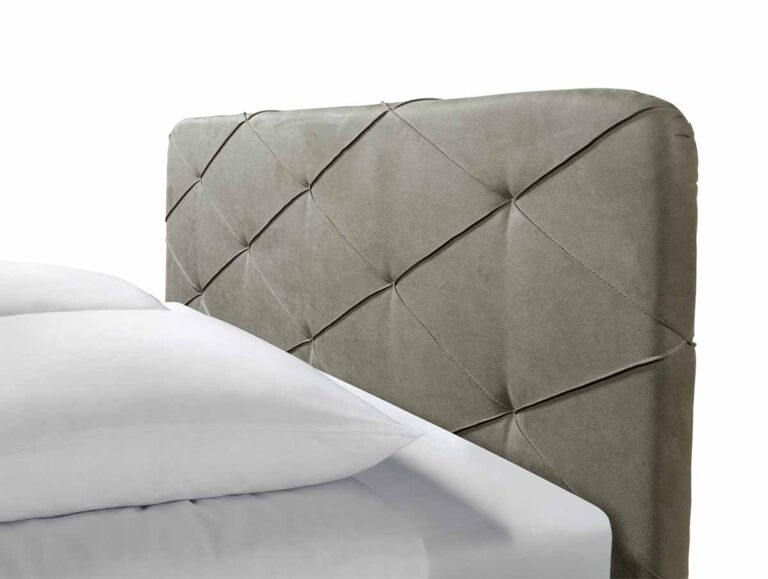 Nahaufnahme eines Bettes mit getuftetem, rautenförmig gemustertem, gepolstertem Kopfteil aus weichem grauem Stoff. Das Bett ist mit weißem Bettzeug und Kissen ausgestattet. Der Hintergrund ist einfarbig weiß.