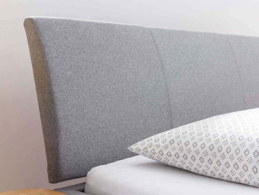 Nahaufnahme eines Bettes mit einem grau gepolsterten Kopfteil, einem weißen Kissen mit geometrischem Muster und einer kleinen grünen Pflanze in einem geflochtenen Topf auf einem Nachttisch mit einer Tasse und Untertasse.