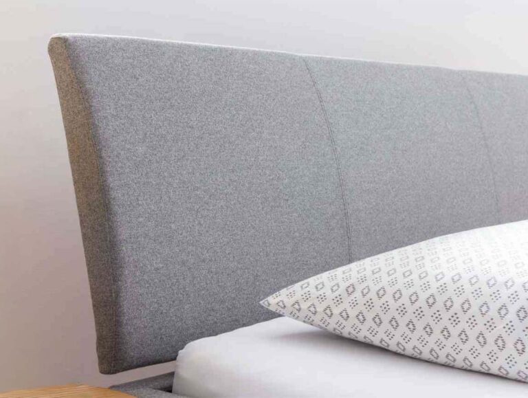 Nahaufnahme eines Bettes mit einem grau gepolsterten Kopfteil, einem weißen Kissen mit geometrischem Muster und einer kleinen grünen Pflanze in einem geflochtenen Topf auf einem Nachttisch mit einer Tasse und Untertasse.