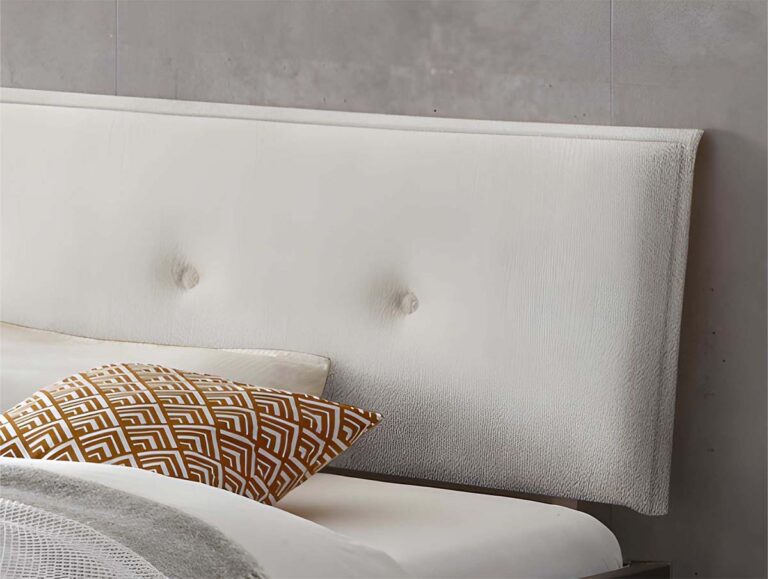 Ein modernes Bett mit einem weiß gepolsterten Kopfteil mit zwei getufteten Knöpfen, einem weißen Laken und einem Zierkissen mit einem geometrischen braun-weißen Muster. Die Wand im Hintergrund ist ein glattes, helles Grau.