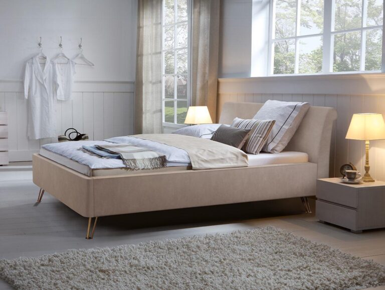 Das Hasena Bett "Dream-Line Ciara 28 Fasio" mit beigen Polstern, Kissen und einer Decke steht in einem hellen Schlafzimmer mit großen Fenstern, hellen Wänden, Nachttischen mit Lampen, einem weißen Teppich und einem Bademantel an der Wand.