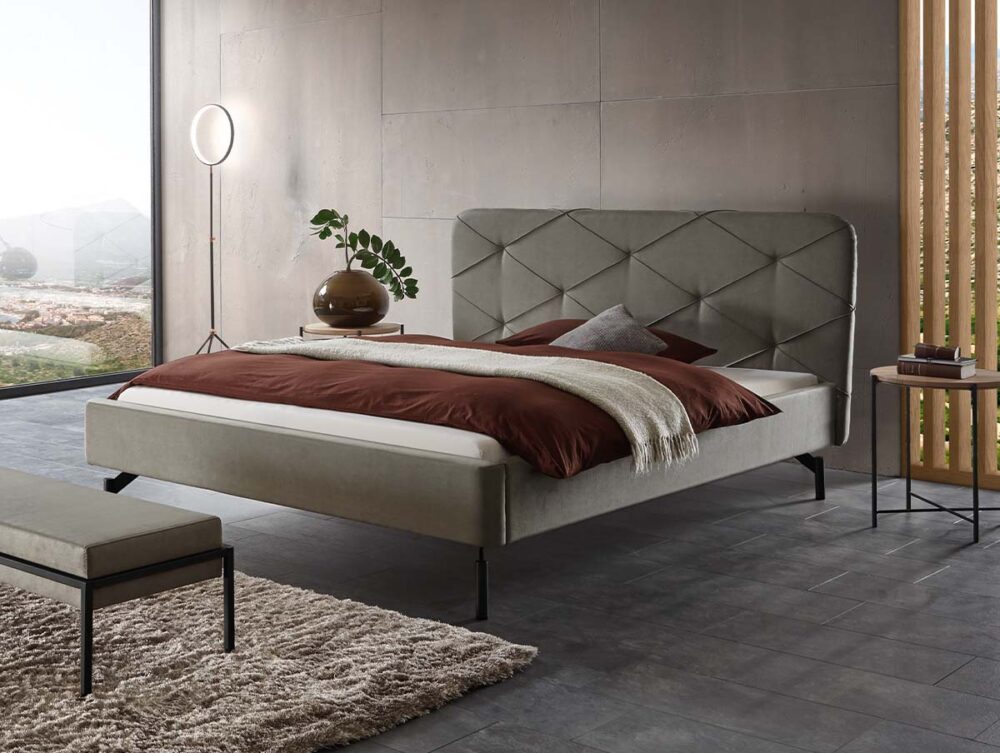 Modernes Schlafzimmer mit einem großen Bett mit grauem Polsterkopfteil, braunem und beigem Bettzeug, einer kleinen Bank, einer Topfpflanze auf dem Nachttisch und großen Fenstern mit Blick ins Freie.