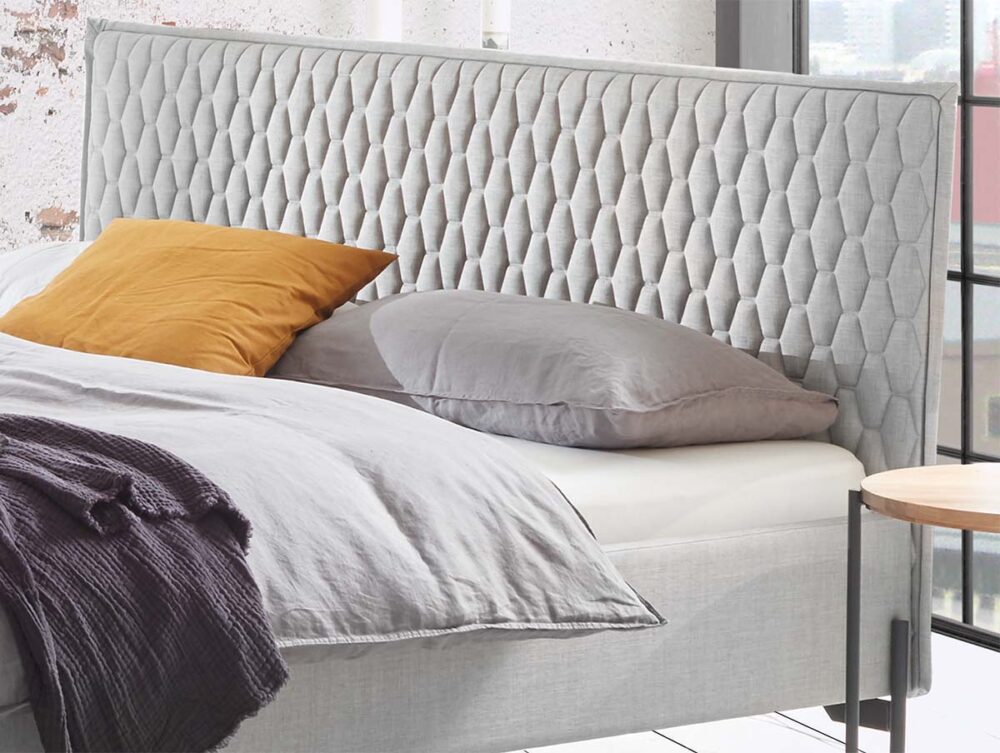 Ein modernes Schlafzimmer mit dem Hasena Polsterbett "Dream-Pronto Gloria" mit einem grauen, getufteten Kopfteil, heller Bettwäsche, teal- und orangefarbenen Kissen, einer dunklen Decke und einem runden Holznachttisch mit Lampe und Klebebandabroller.