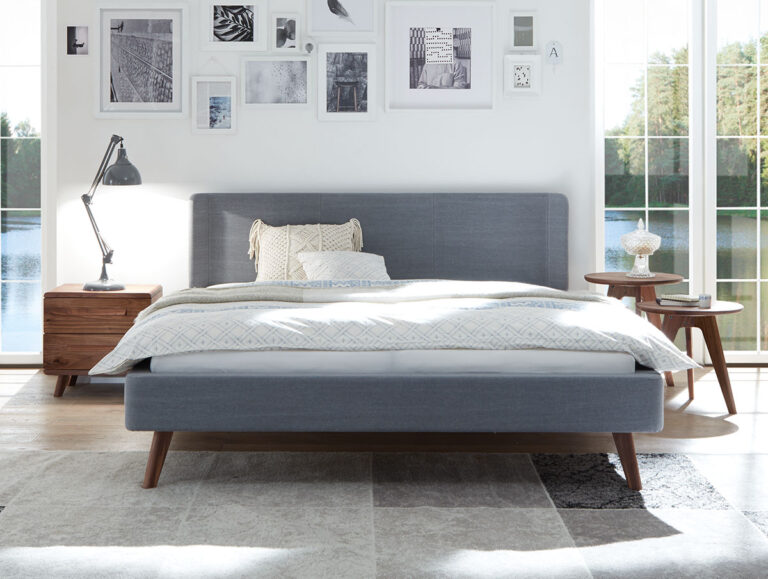 Ein modernes Schlafzimmer mit einem grauen Polsterbett, weißem Bettzeug, Nachttischen aus Holz auf beiden Seiten, einer Tischlampe und gerahmter Kunst an der weißen Wand. Große Fenster zeigen Bäume und einen See draußen.