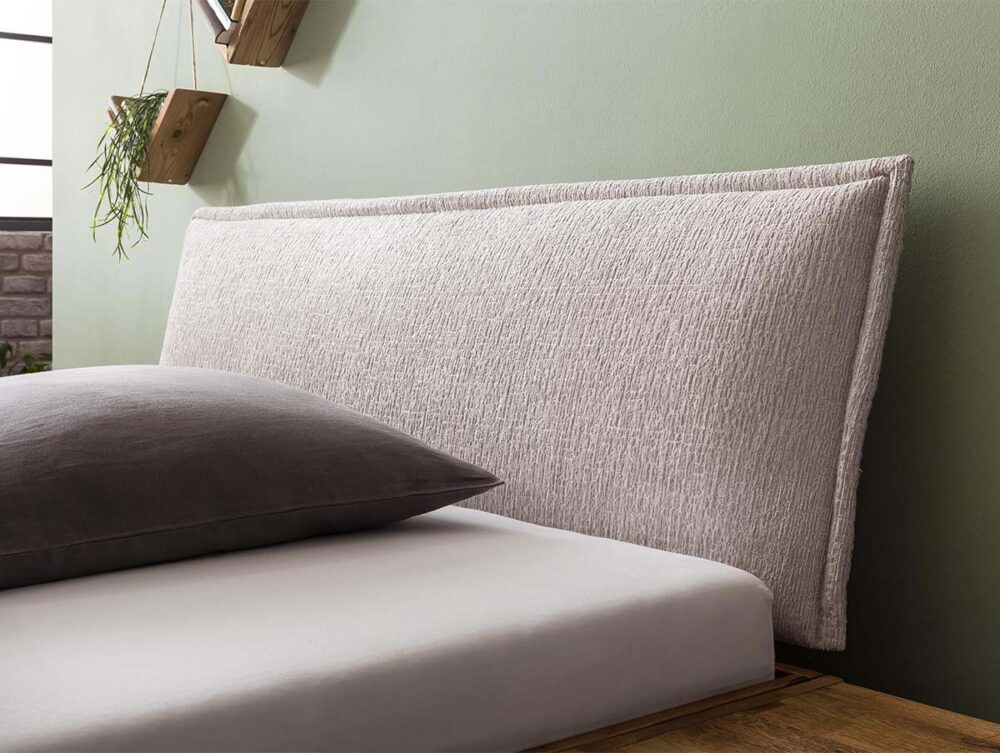 Ein modernes Bett mit einem strukturierten, hellbeigen Polsterkopfteil, einem grauen Kissen und einem weißen Laken. Das Zimmer hat hellgrüne Wände und Holzregale mit Topfpflanzen.