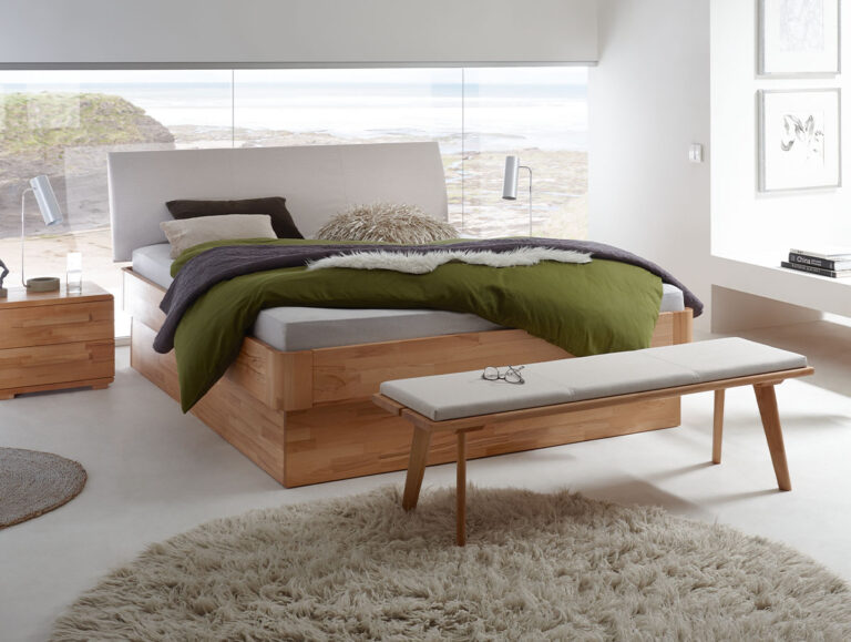 Ein modernes Schlafzimmer zeigt die "Fine-Line Syma Practico-Ron Box 1" von Hasena Bett, die mit grüner und weißer Bettwäsche, einer Bank am Fußende, einem runden beigen Teppich, großen Fenstern mit Panoramablick, minimalistischem Dekor und weichem natürlichem Licht ausgestattet ist.