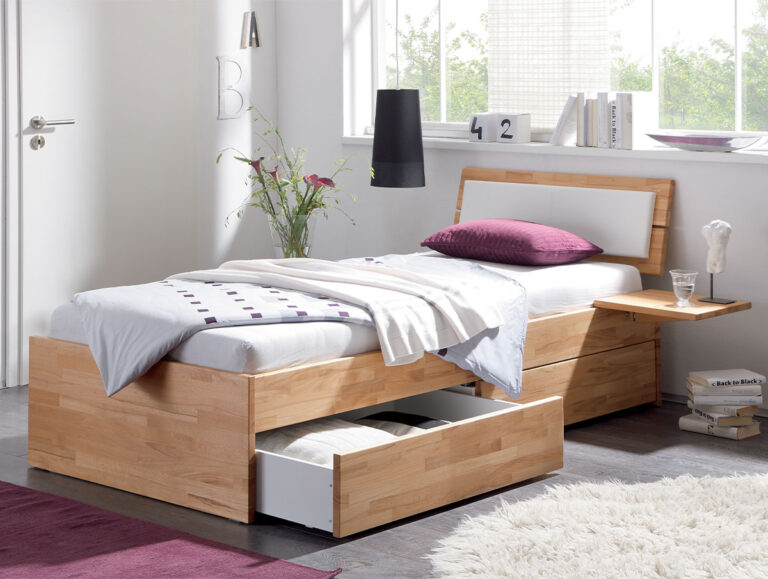 Ein modernes Hasena Schubladenbett "Function and Comfort Spazio 2" mit weißer Bettwäsche, lilafarbenem Kissen und Schubladen darunter. Ein Seitenregal bietet Platz für ein Glas und Bücher; Blumen, ein Teppich und natürliches Licht bereichern das minimalistische Zimmer.