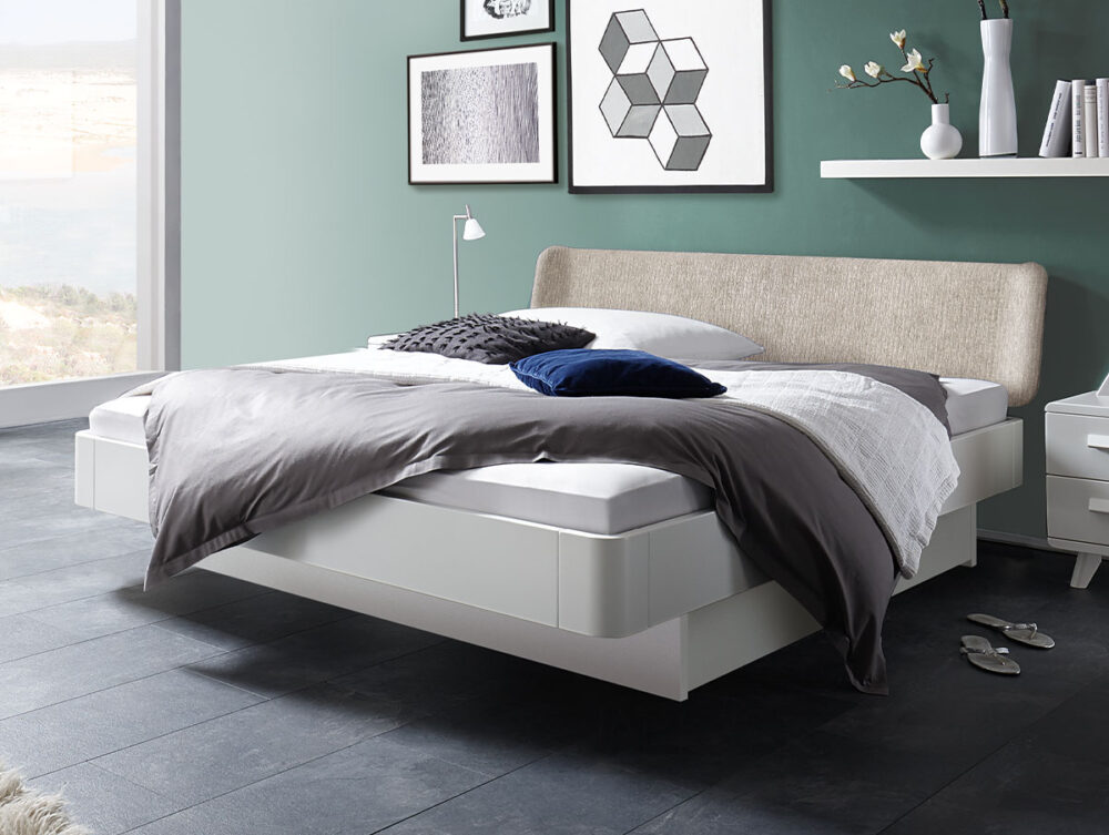 Ein modernes Schlafzimmer mit einem weißen Schwebebett, grauer und weißer Bettwäsche und dunklen Bodenfliesen. Die Wand ist grün und mit gerahmter Kunst verziert. Ein kleiner Nachttisch, eine Lampe und ein Regal mit Vasen stehen neben dem Bett.