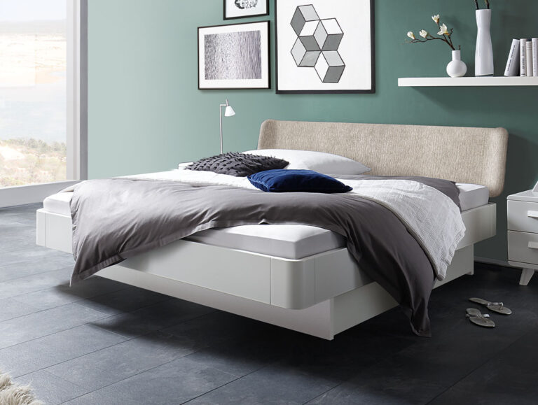 Ein modernes Schlafzimmer mit einem weißen Schwebebett, grauer und weißer Bettwäsche und dunklen Bodenfliesen. Die Wand ist grün und mit gerahmter Kunst verziert. Ein kleiner Nachttisch, eine Lampe und ein Regal mit Vasen stehen neben dem Bett.