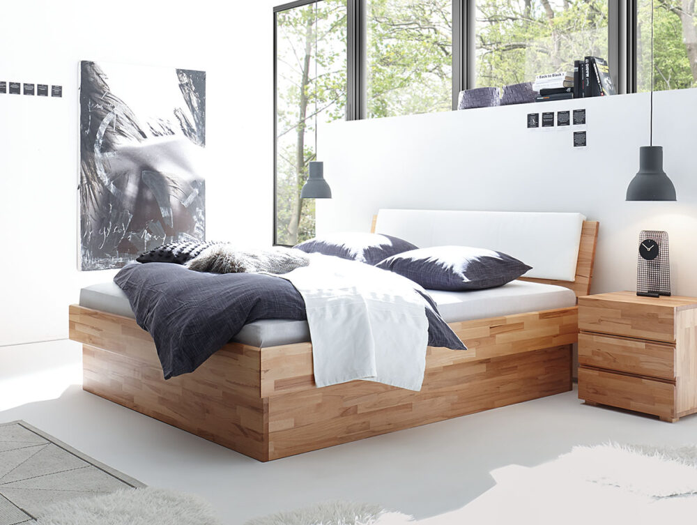 Ein modernes Schlafzimmer mit dem Hasena Bett "Wood-Line Premium 18 Practico-Box" Bettrahmen, weißer und dunkler Bettwäsche, einem hölzernen Nachttisch mit Uhr, großen Fenstern, schwarzen Hängelampen, einem zotteligen weißen Teppich und schwarz-weißer abstrakter Wandkunst.