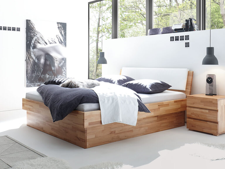 Ein modernes Schlafzimmer mit dem Hasena Bett "Wood-Line Premium 18 Practico-Box" Bettrahmen, weißer und dunkler Bettwäsche, einem hölzernen Nachttisch mit Uhr, großen Fenstern, schwarzen Hängelampen, einem zotteligen weißen Teppich und schwarz-weißer abstrakter Wandkunst.