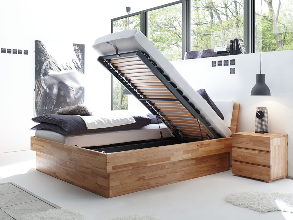 Das Hasena Bett "Wood-Line Premium 18 Practico-Box" ist ein Stauraumbett aus Holz mit einer hochklappbaren Matratze für versteckten Stauraum, das neben einem passenden Nachttisch in einem hellen, minimalistischen Schlafzimmer mit großen Fenstern gezeigt wird.