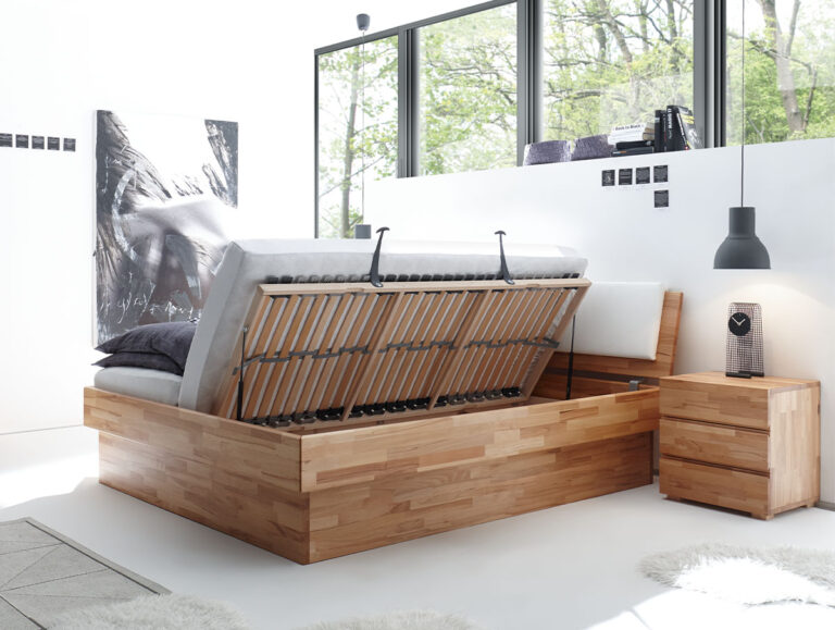 Ein modernes Schlafzimmer zeigt das Hasena Bett "Wood-Line Premium 18 Practico-Box" Stauraumbett, dessen Matratze angehoben wird, um versteckten Stauraum freizugeben, ergänzt durch weiße Wände, große Fenster, einen Nachttisch und abstrakte Kunst im natürlichen Licht.