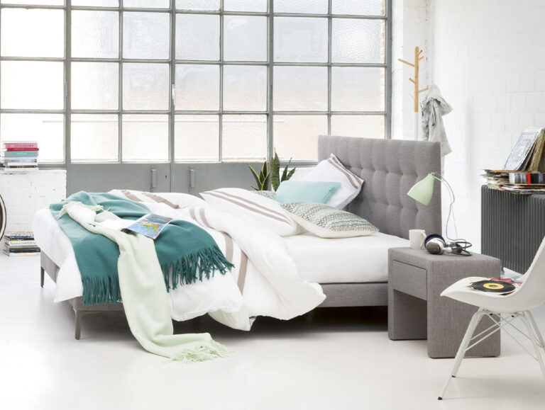 In einem hellen, modernen Loft mit großen Fenstern stehen ein Bett mit türkisfarbener und weißer Bettwäsche, ein Fahrrad, ein Tisch, an dem eine Person schreibt, ein weißer Stuhl und verschiedene Dekorationen, die eine gemütliche, wohnliche Atmosphäre schaffen.