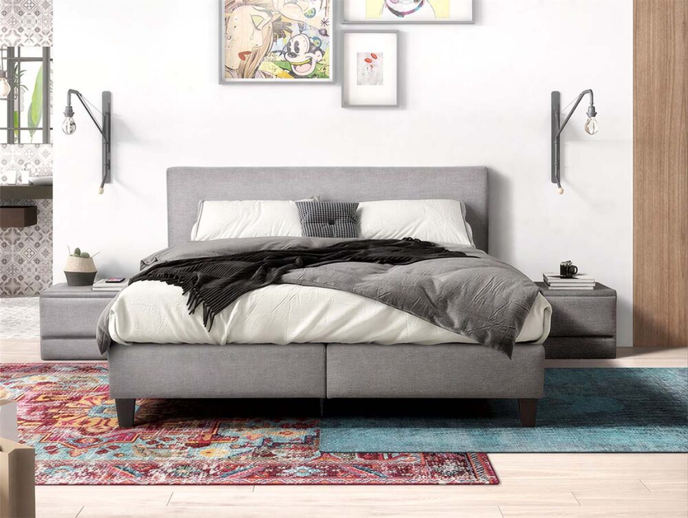 Ein modernes Schlafzimmer mit einem grauen Polsterbett, weißer Bettwäsche und einer dunklen Überwurfdecke. An den Wänden sind auf jeder Seite Lampen angebracht, auf dem Boden liegen bunte Teppiche und an der weißen Wand über dem Bett hängt gerahmte Kunst.