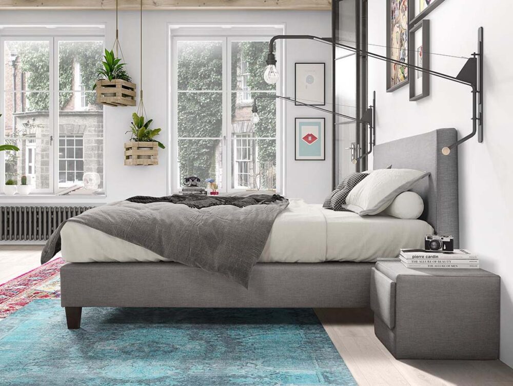 Ein modernes Schlafzimmer mit einem grauen Polsterbett, weißer und grauer Bettwäsche, einer Wandlampe, schwebenden Pflanzen, großen Fenstern und einem blauen Teppich auf hellem Holzfußboden. Gerahmte Kunstwerke schmücken die Wand über dem Bett.