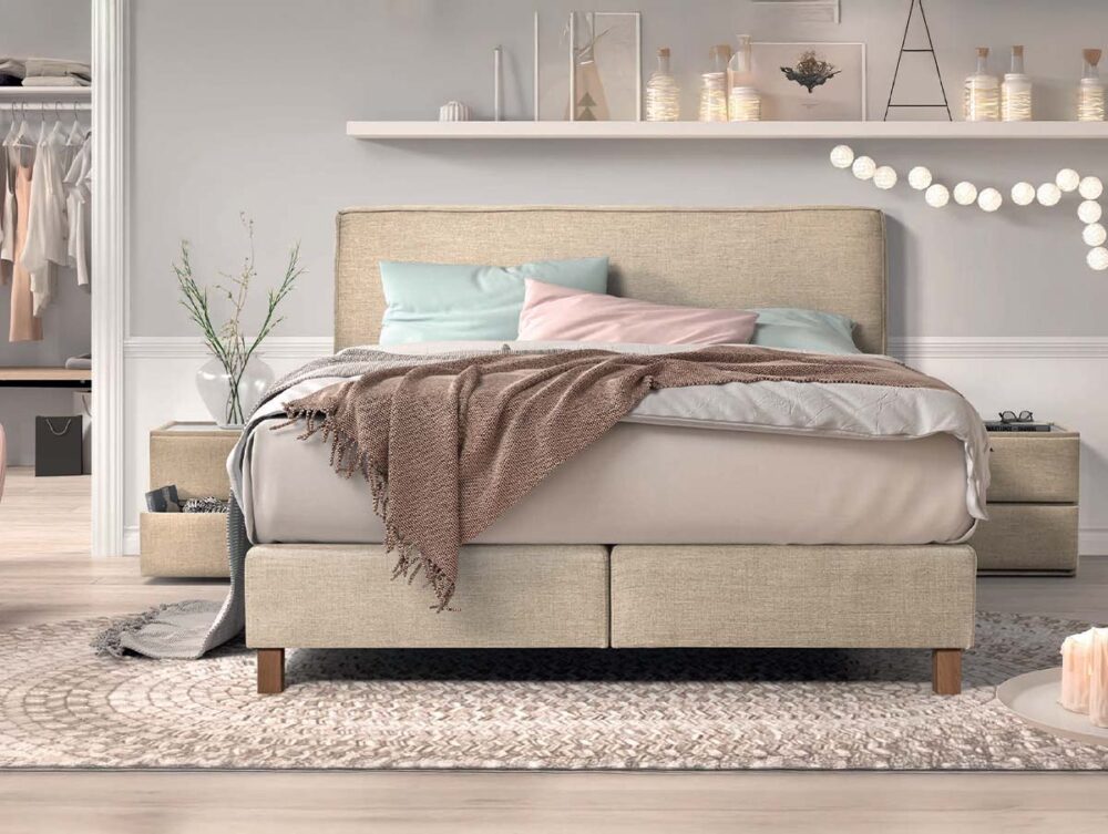 Ein gemütliches, modernes Schlafzimmer mit einem beigen Bett, pastellfarbenen Kissen und einer rosa Decke. Kunstdrucke, Kerzen und Lichterketten schmücken das Wandregal. Auf der linken Seite sind ein rosafarbener Sessel und ein offener begehbarer Kleiderschrank zu sehen.