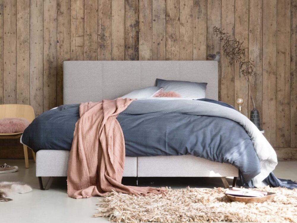 Ein gemütliches Schlafzimmer mit einem grauen Polsterbett, blauer Bettwäsche und einer rosa Überwurfdecke. Das Zimmer verfügt über eine Wand aus Holzdielen, einen beigen Teppich, einen einfachen Stuhl mit einem rosa Kissen und einen kleinen Nachttisch mit Dekoration.