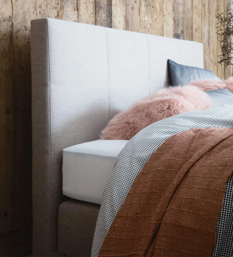 Ein modernes Bett mit einem hellgrauen, gepolsterten Kopfteil, das mit weißen Laken, einer rosa Kunstpelzdecke, einem grauen Kissen und einer rostfarbenen, strukturierten Decke vor einer rustikalen Holzwand sauber bezogen ist.