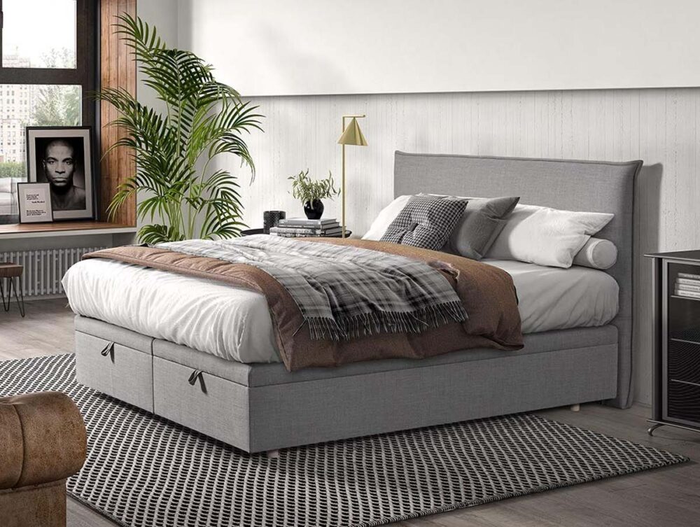 Ein modernes Schlafzimmer mit einem grauen Polsterbett, weißer Bettwäsche, einer karierten Decke, verschiedenen Kissen, einem gemusterten Teppich, einer Topfpflanze, gerahmten Fotos und einem großen Fenster, das natürliches Licht hereinlässt.