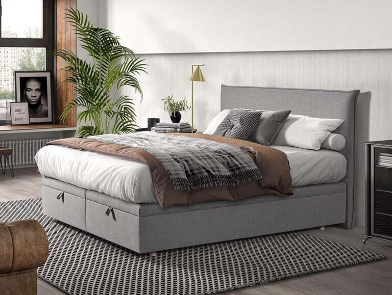 Ein modernes Schlafzimmer mit einem grauen Polsterbett, weißer Bettwäsche, einer karierten Decke, verschiedenen Kissen, einem gemusterten Teppich, einer Topfpflanze, gerahmten Fotos und einem großen Fenster, das natürliches Licht hereinlässt.