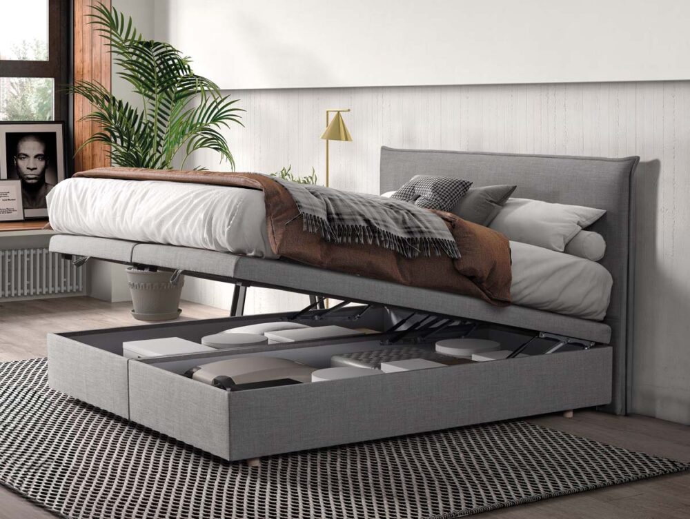 Ein graues gepolstertes Bett, dessen Matratze hochgeklappt ist, so dass darunter ein Stauraum mit Kisten zum Vorschein kommt. Im Zimmer stehen eine Pflanze, ein gerahmtes Foto, ein Fenster mit Sonnenlicht und eine neutrale Einrichtung.