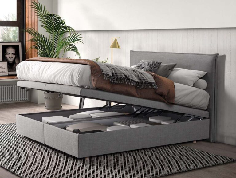 Ein graues gepolstertes Bett, dessen Matratze hochgeklappt ist, so dass darunter ein Stauraum mit Kisten zum Vorschein kommt. Im Zimmer stehen eine Pflanze, ein gerahmtes Foto, ein Fenster mit Sonnenlicht und eine neutrale Einrichtung.