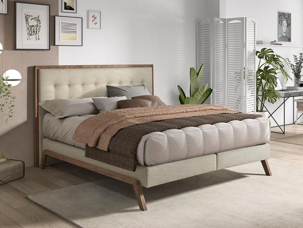 Ein modernes Schlafzimmer mit einem beigen Polsterbett, neutraler Bettwäsche und einer braunen Decke. Das Zimmer hat gerahmte Kunstwerke an der Wand, Pflanzen, einen Raumteiler und einen Schreibtisch in der Ecke, alles auf hellem Holzboden mit einem beigen Teppich.