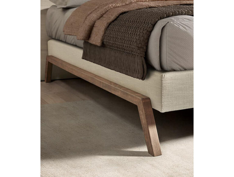 Nahaufnahme eines beigefarbenen, gepolsterten Bettgestells mit abgewinkelten Holzbeinen, darauf ordentlich geschichtete taupefarbene und braune Decken, auf einem hellen Teppich und Holzboden.