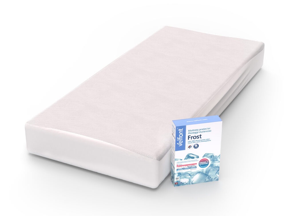 Eine weiße Matratze mit einem hellen Bezug ist hinter einem Karton mit der Aufschrift Velfont Fixleintuch Thermoregulierend angebracht. Auf der Schachtel sind Eiswürfelbilder und Produktdetails abgebildet, die die thermoregulierenden Eigenschaften dieser Unterlage für optimalen Komfort hervorheben.