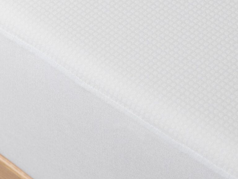 Nahaufnahme der Velfont Fixleintuch Thermoregulierend Matratze mit weißem Bezug und dezentem Gittermuster auf einem Holzbettgestell.