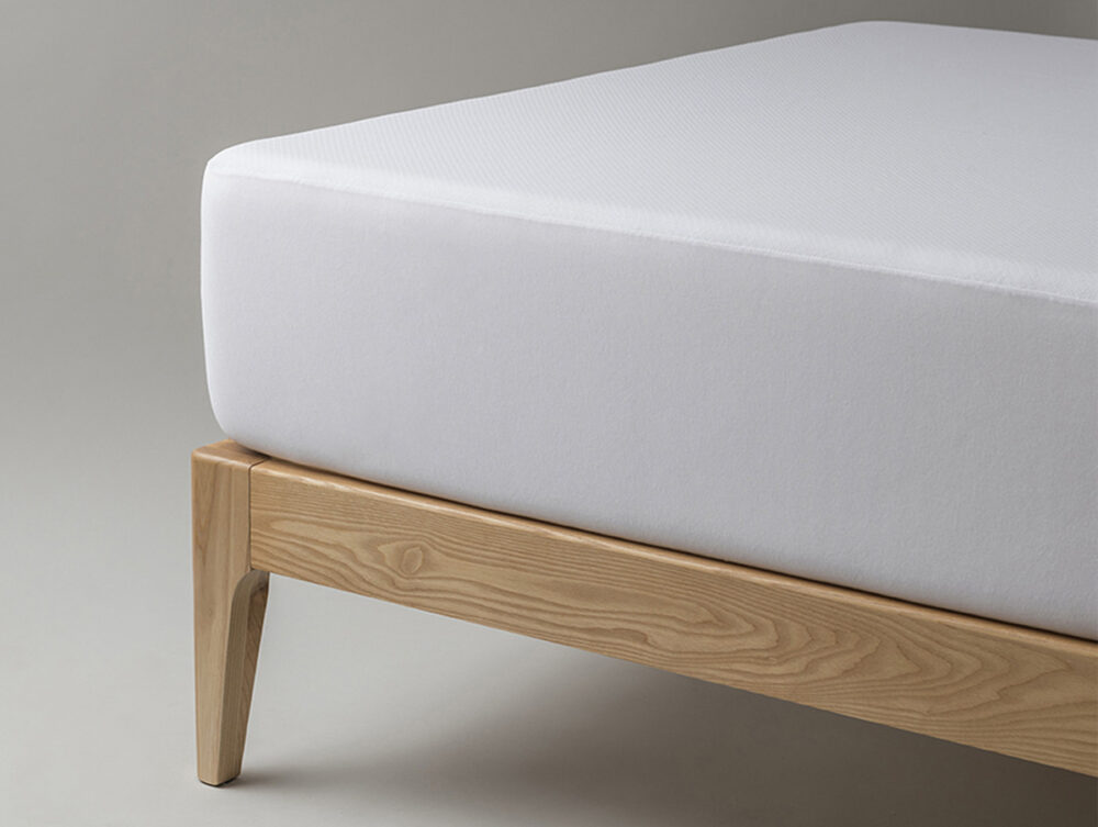 Nahaufnahme einer Bettecke mit hellem Holzrahmen, auf der das Velfont Fixleintuch Thermoregulierend in Weiß auf der Matratze liegt, vor einem einfarbig grauen Hintergrund.