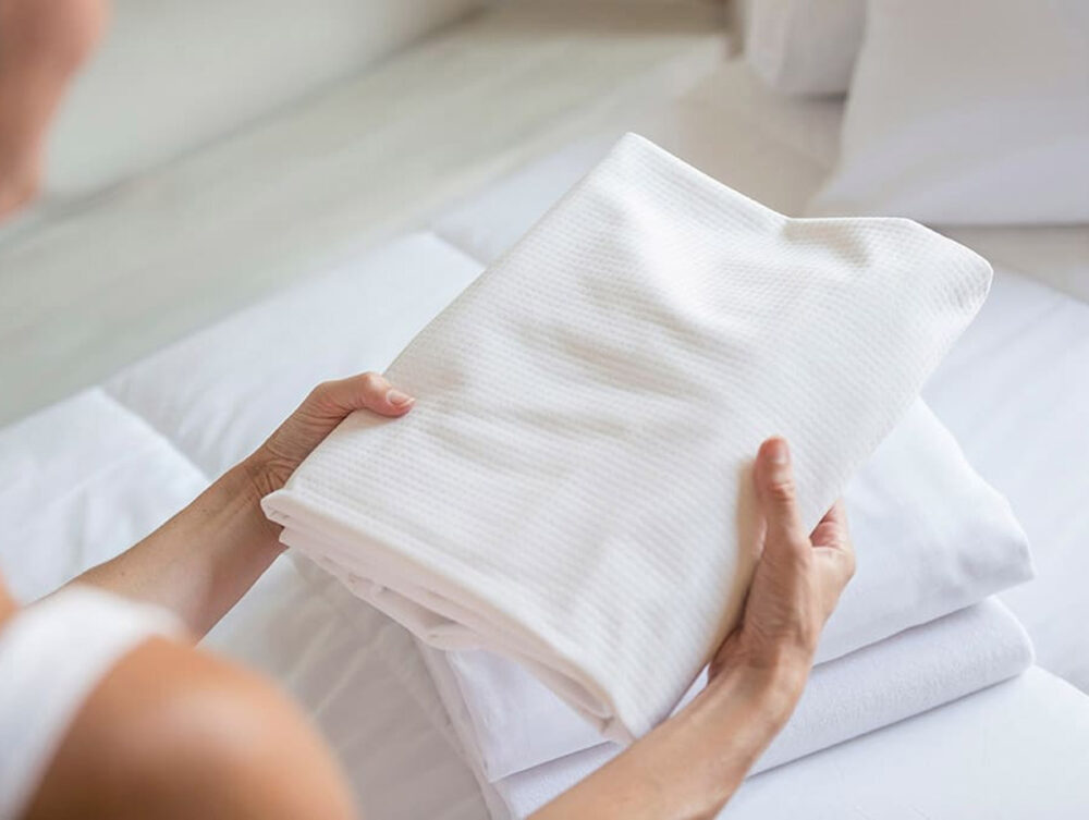 Eine Person hält ein ordentlich gefaltetes weißes Handtuch über einen Stapel Velfont Fixleintuch Thermoregulierend auf einem Bett in einem hellen, sauberen Raum.