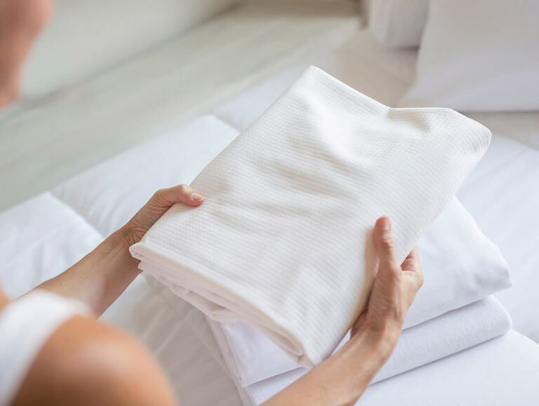 Eine Person hält ein ordentlich gefaltetes weißes Handtuch über einen Stapel Velfont Fixleintuch Thermoregulierend auf einem Bett in einem hellen, sauberen Raum.