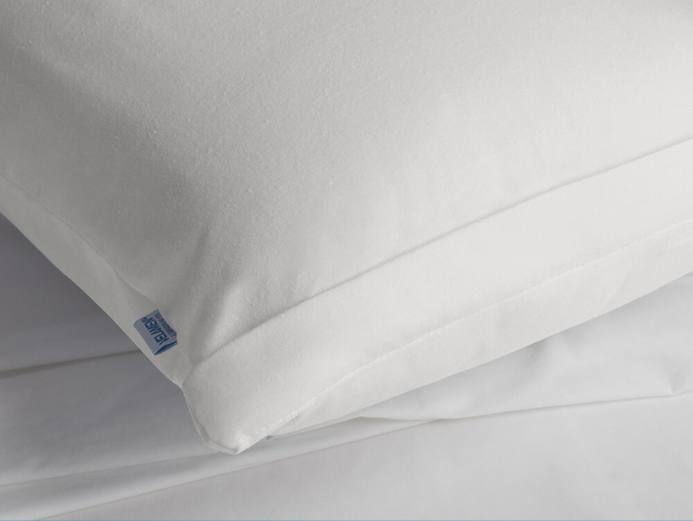 Ein strahlend weißes Kissen mit einem blauen Etikett ruht auf einem glatten weißen Laken und präsentiert den Velfont Kissenbezug Respira wasserdicht für hervorragenden Schutz und Komfort.