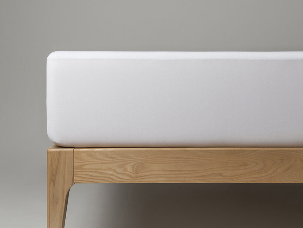 Ein minimalistisches Bettgestell aus Holz mit einer dicken, weißen Matratze darauf vor einem schlichten grauen Hintergrund.