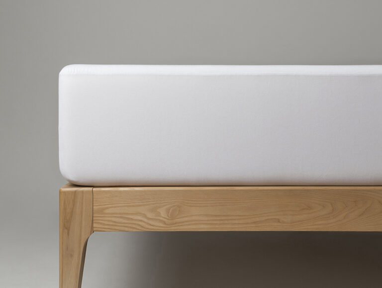 Ein minimalistisches Bettgestell aus Holz mit einer dicken, weißen Matratze darauf vor einem schlichten grauen Hintergrund.