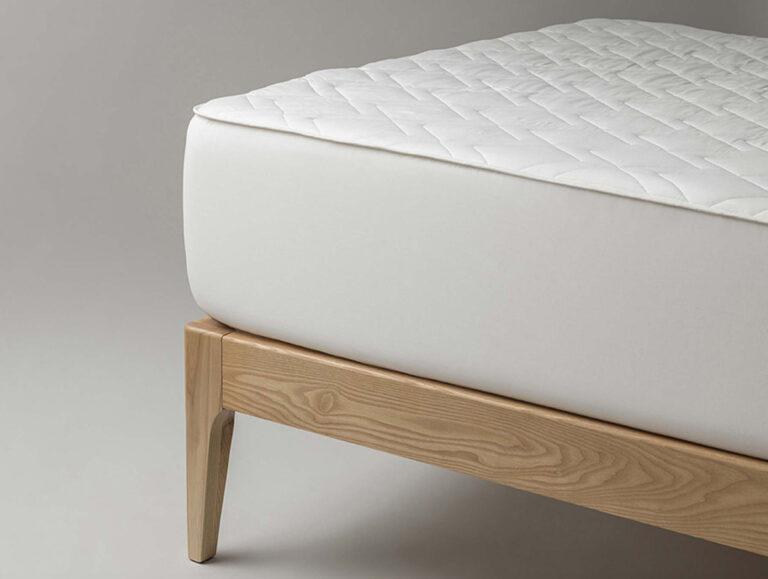 Eine Nahaufnahme einer sauber gefertigten Matratze mit weißem Steppbezug auf einem hellen Holzbettgestell vor einem schlichten grauen Hintergrund.