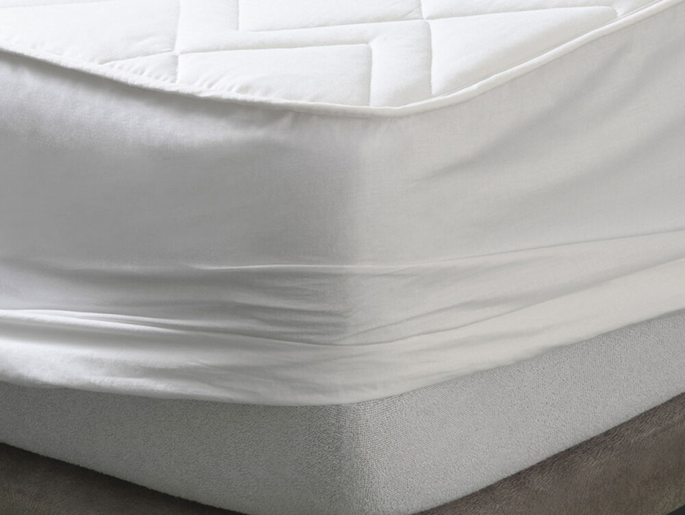 Eine Nahaufnahme einer Matratze mit einem weißen Spannbettlaken und einem gesteppten Matratzenschoner darauf, der glatte, sauber umwickelte Ecken und sauberen Stoff zeigt.