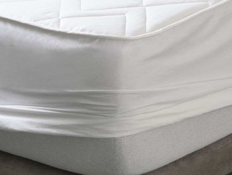 Eine Nahaufnahme einer Matratze mit einem weißen Spannbettlaken und einem gesteppten Matratzenschoner darauf, der glatte, sauber umwickelte Ecken und sauberen Stoff zeigt.