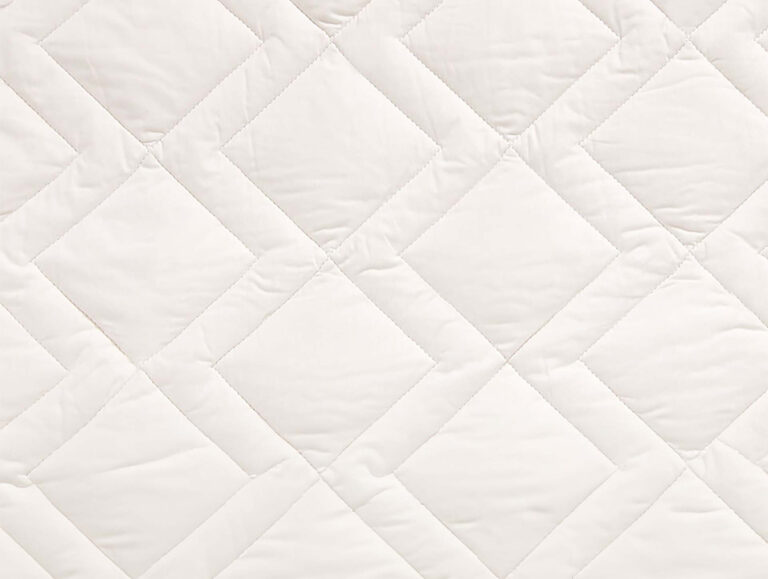 Nahaufnahme eines gesteppten Stoffes mit Rautenmuster, der weiße Nähte und eine weiche, gepolsterte Textur aufweist. Die Oberfläche erscheint glatt und sauber, mit leichten Falten und Nähten zwischen den Rautenformen.