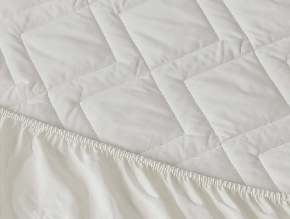 Nahaufnahme eines weißen, gesteppten Matratzenschoners mit Rautensteppung und elastischem Rand, der eine Matratze teilweise bedeckt.