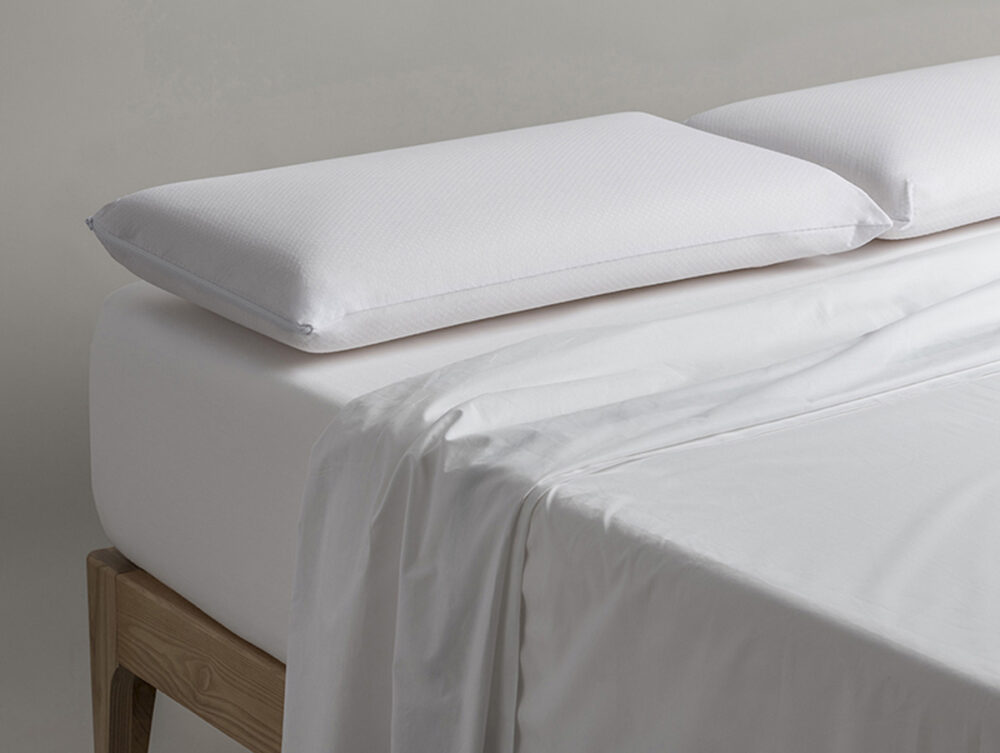 Ein sauber gemachtes Bett mit Holzrahmen ist mit einem weißen Spannbettlaken, einem teilweise zurückgezogenen flachen Laken und dem Velfont Kopfkissen "Viscofresh" Thermoregulierend vor einem schlichten Hintergrund ausgestattet.