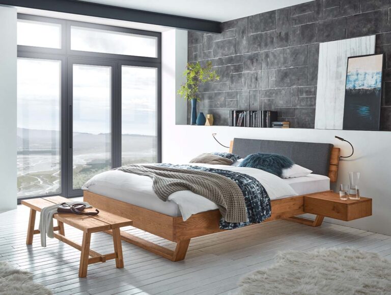 Ein modernes Schlafzimmer zeigt das Hasena Bett "Oak-Wild Cadro 18 Skid", gepaart mit einer Holzbank, einem grauen Kopfteil, blauer und grauer Bettwäsche, einem großen Panoramafenster, einer grau gefliesten Akzentwand, abstrakter Kunst und einer Vase mit Grünzeug auf einem schwebenden Nachttisch.