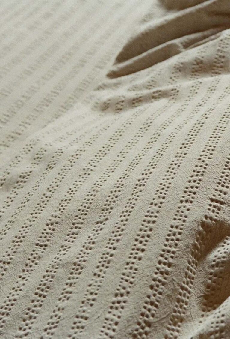 Nahaufnahme eines beigen Stoffes mit kleinen, gleichmäßig verteilten Löchern, die vertikale gepunktete Linien bilden und eine strukturierte und gemusterte Oberfläche erzeugen. Der Stoff scheint sanfte Falten oder Wellen zu haben.