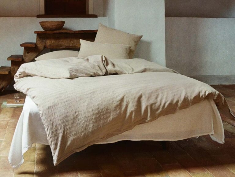 Ein gemütliches, ungemachtes Bett mit beiger Bettwäsche und Kissen steht in einem rustikalen Raum mit einer Holzbank, einer Keramikschüssel und einem gefliesten Boden. Weiches natürliches Licht erfüllt den Raum und schafft eine warme, einladende Atmosphäre.