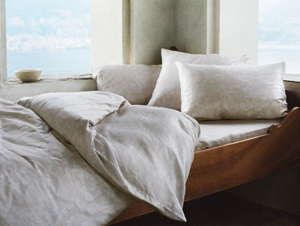 Ein gemütliches Bett mit weicher, heller Bettwäsche und Kissen steht in der Nähe eines Fensters mit Blick auf die Landschaft. Eine kleine Schale steht auf der Fensterbank, und natürliches Licht erhellt den friedlichen, minimalistischen Raum.